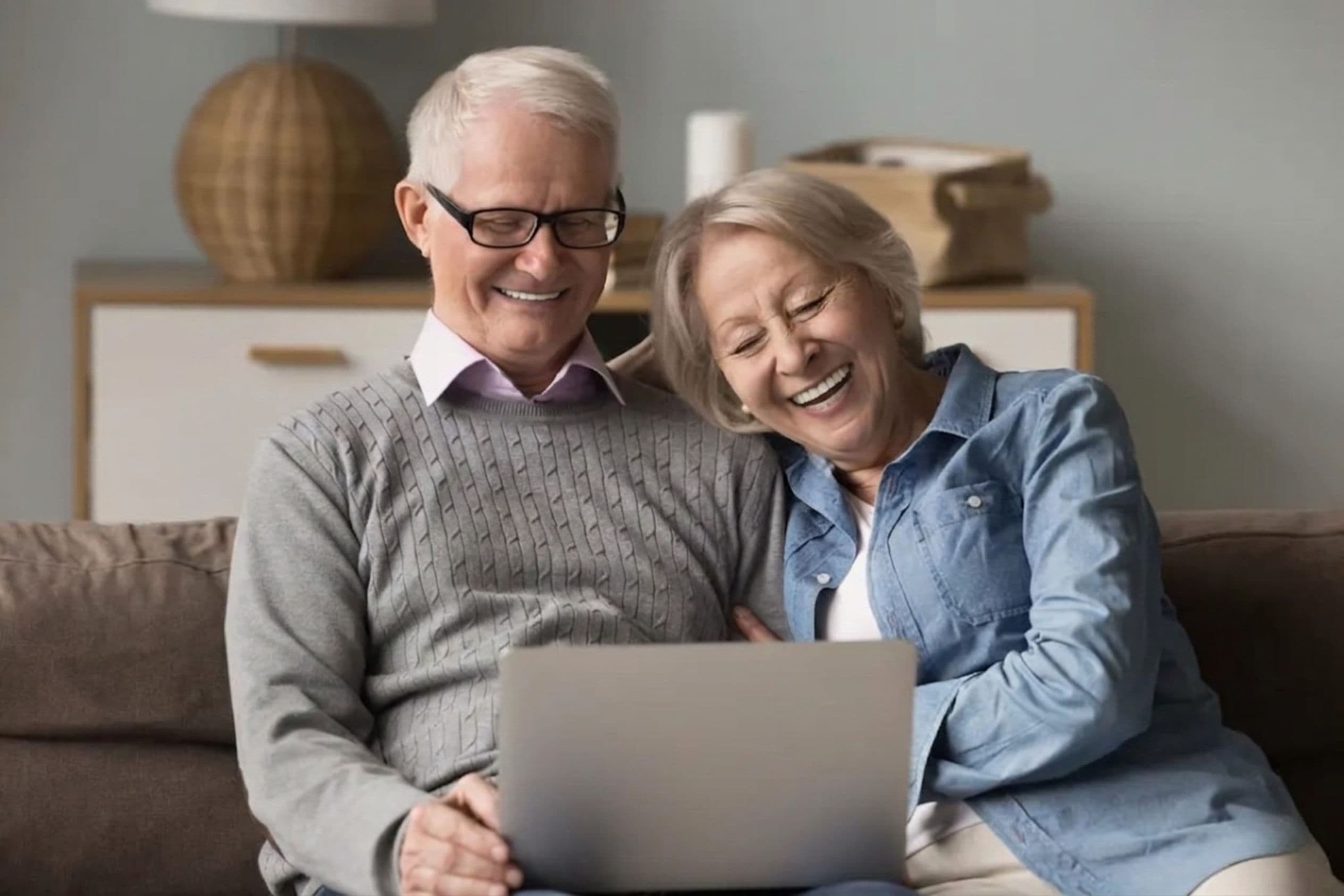 A happy older couple sitting on their sofa at home, laughing together as they look at a laptop